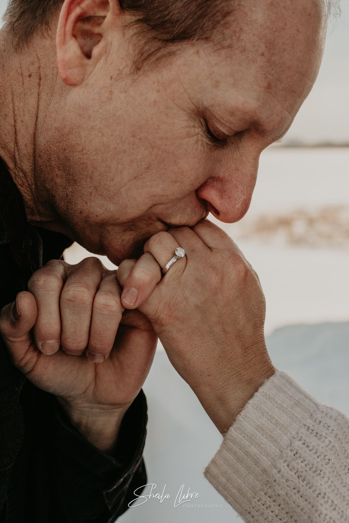 Intimate close-up of gentle hand kiss showing engagement ring in moody lighting.
