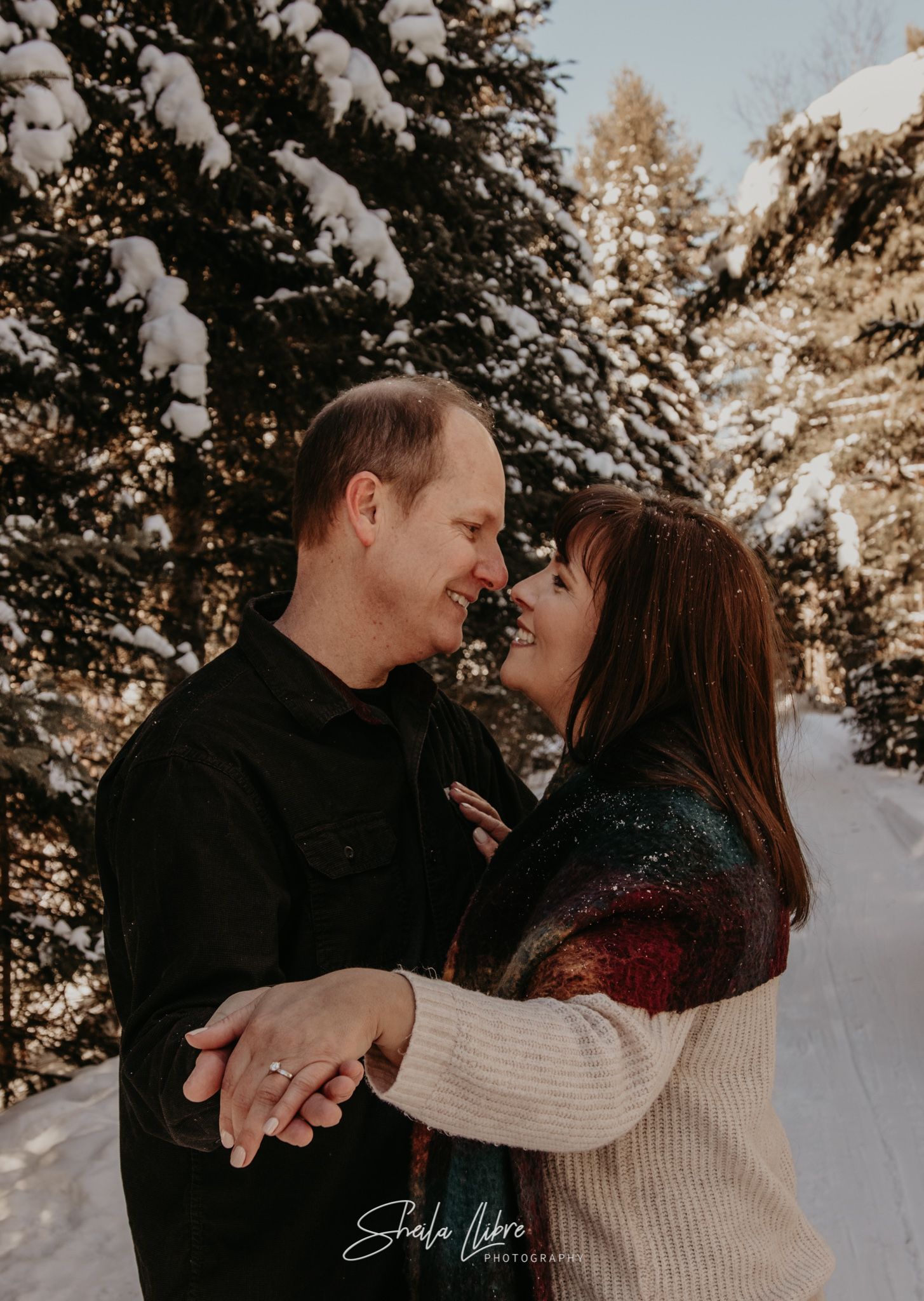 Couple embracing and smiling in snow-covered winter forest setting.