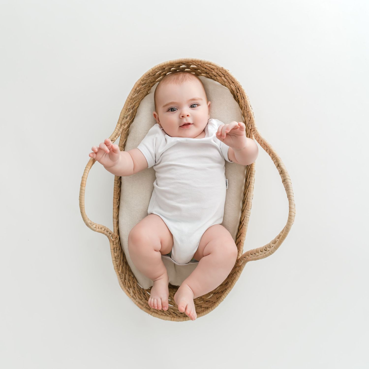 Infant in white onesie lying in woven basket against white background.