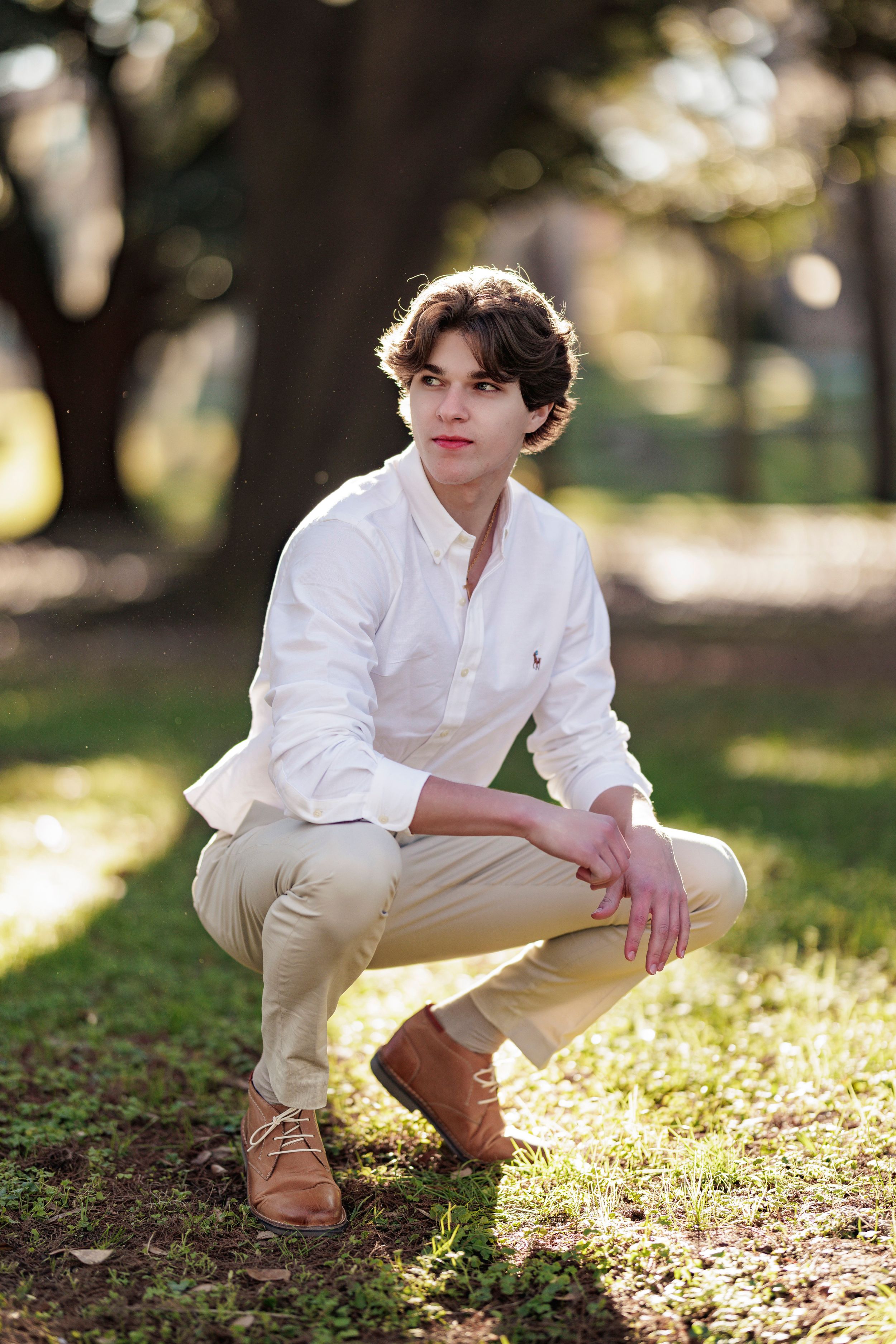 Person in preppy attire crouching in a park setting with dappled sunlight.