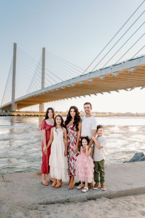 A family poses together on a beach with a large cable-stayed bridge visible in the background at sunset.