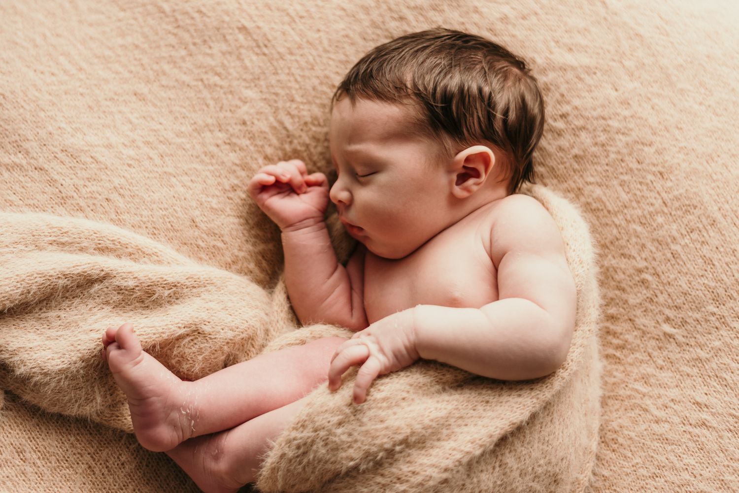 Newborn baby sleeping peacefully on beige blanket with hand near face.