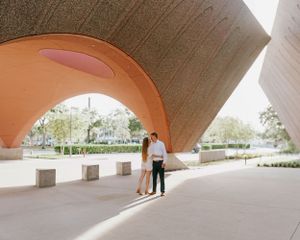 Couple standing under a large curved orange architectural structure.