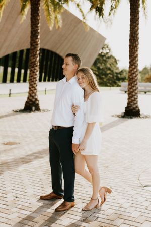 A couple in matching white outfits standing together on a palm tree-lined brick pathway.