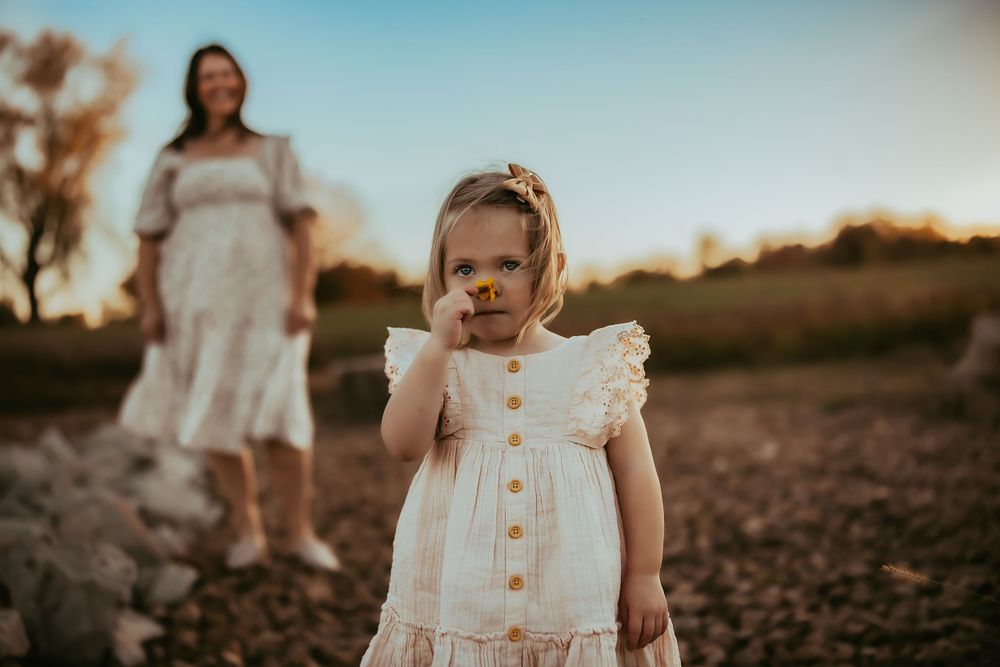 Little girl smelling a flower with mom watching in the distance.