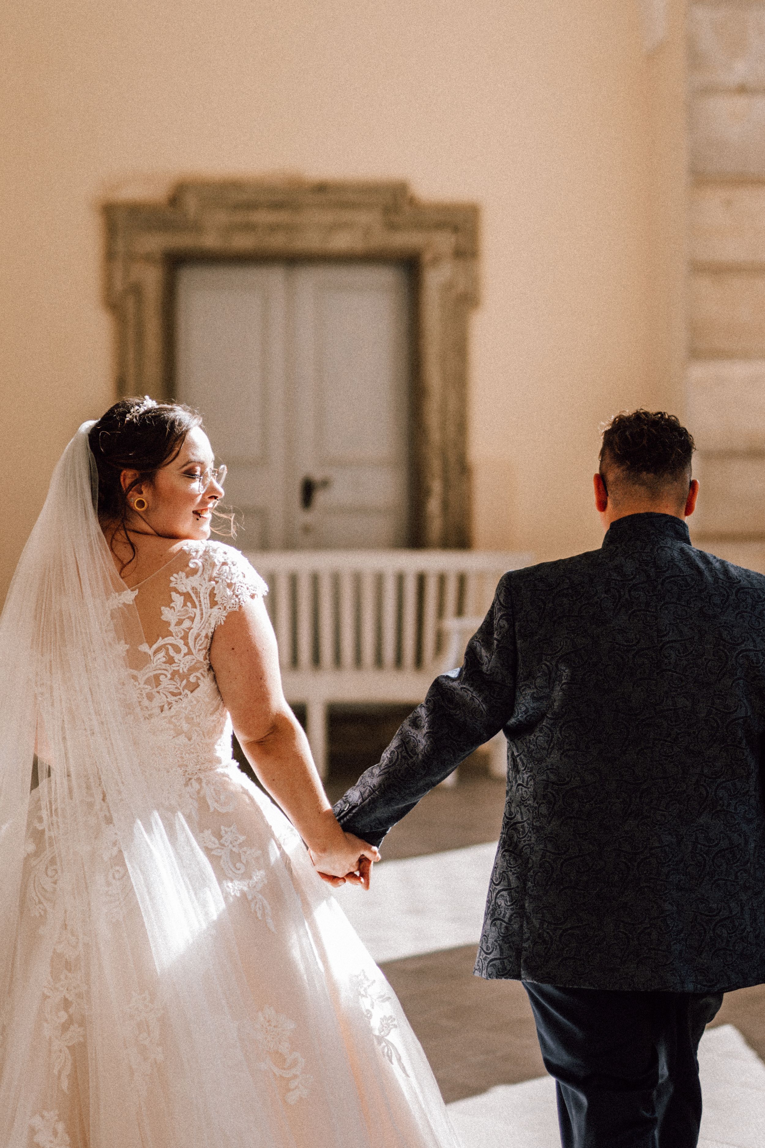 A romantic wedding moment as a couple holds hands walking through an elegant hallway with ornate decor.