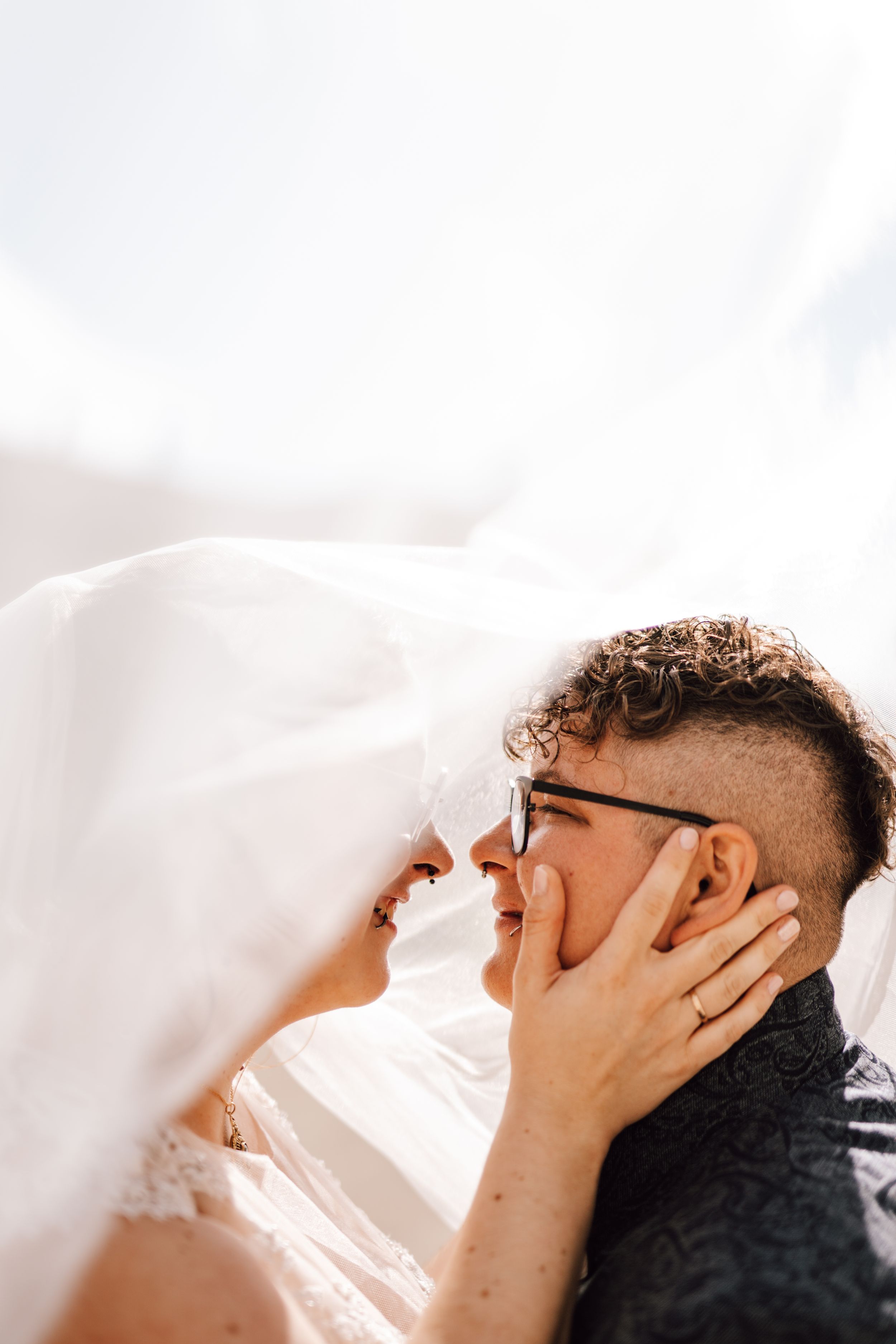 An intimate wedding moment captured under a flowing white veil against a bright background.