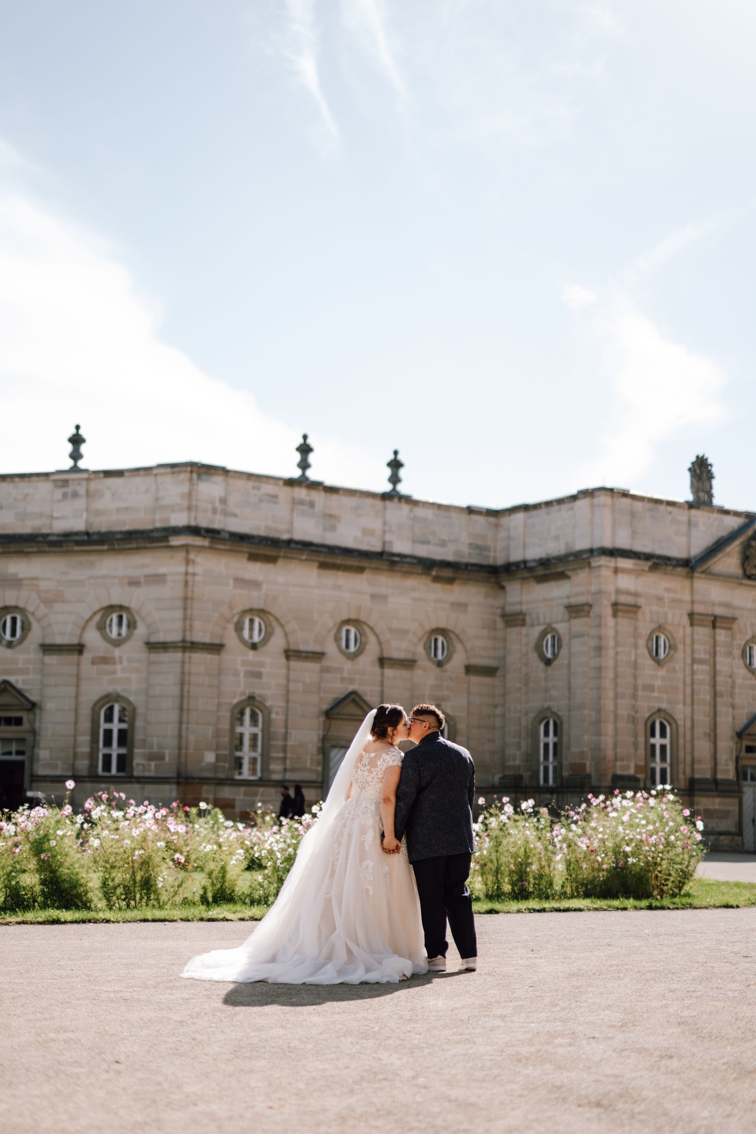 A newlywed couple stands together in front of a grand historic building with blooming flowers.