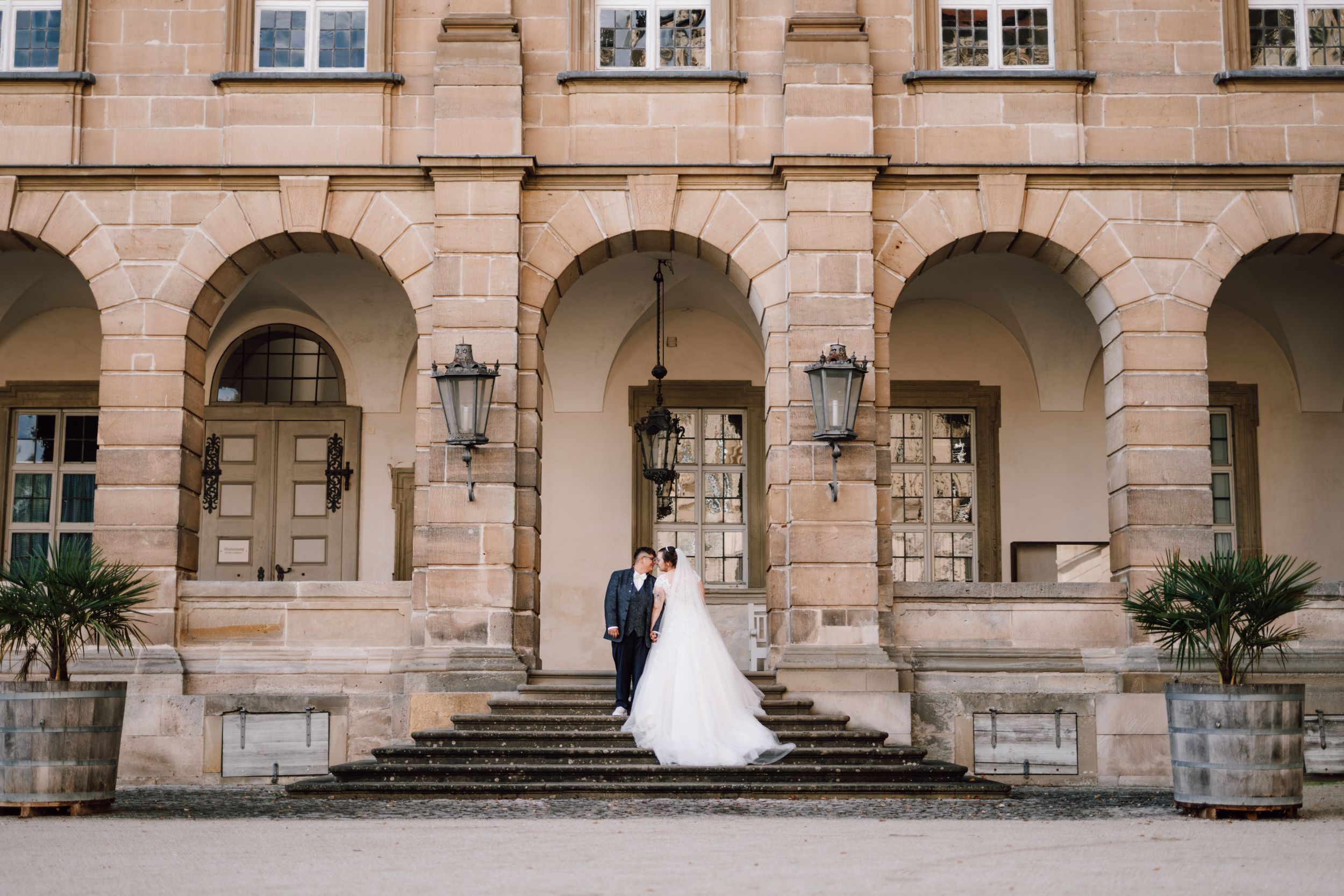 A wedding couple poses on grand stone steps of a classical architecture building with arches.