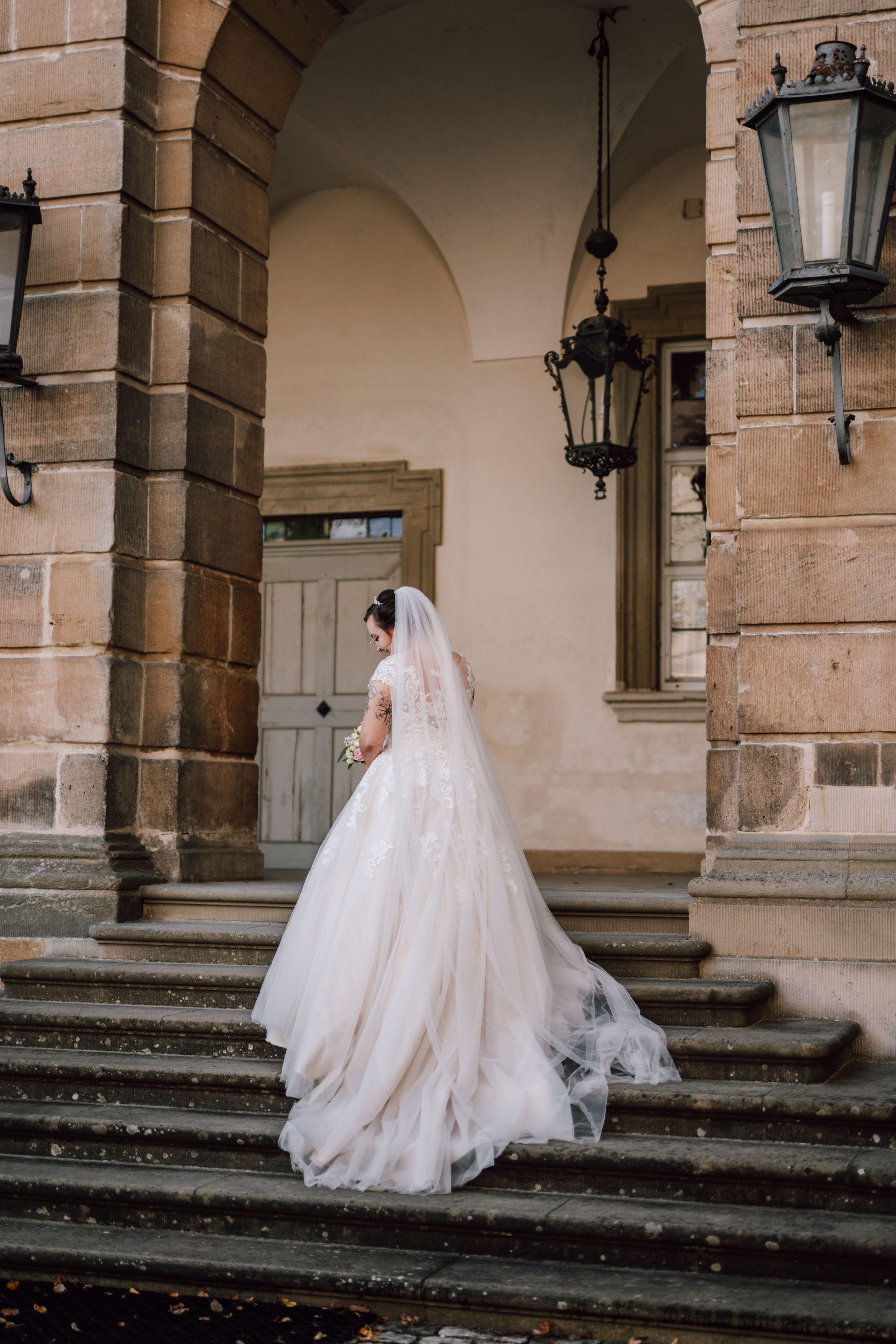 A bride in a flowing white gown ascends stone steps at a historic venue with ornate lanterns.