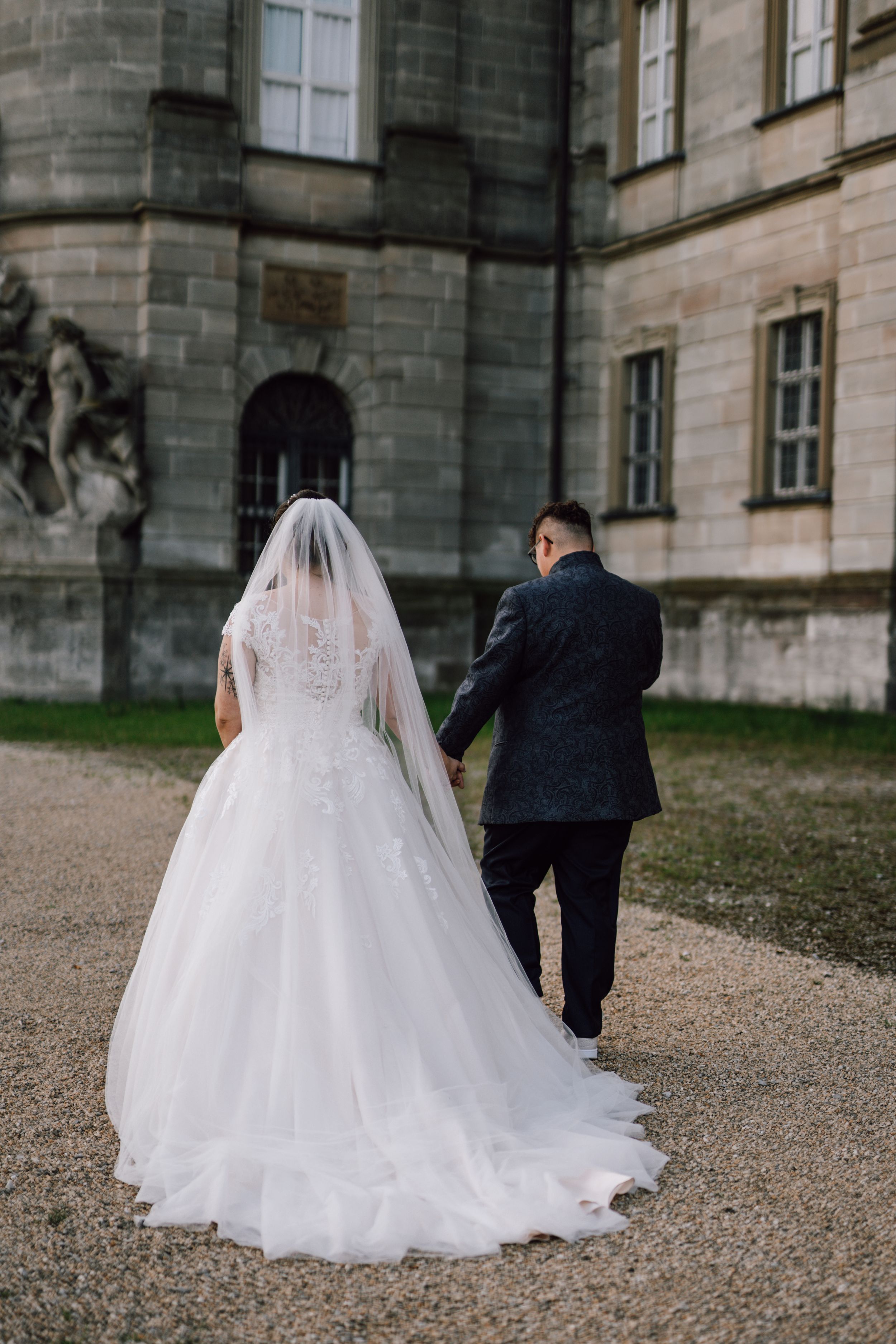 A wedding couple photographed from behind walking near a historic stone building.