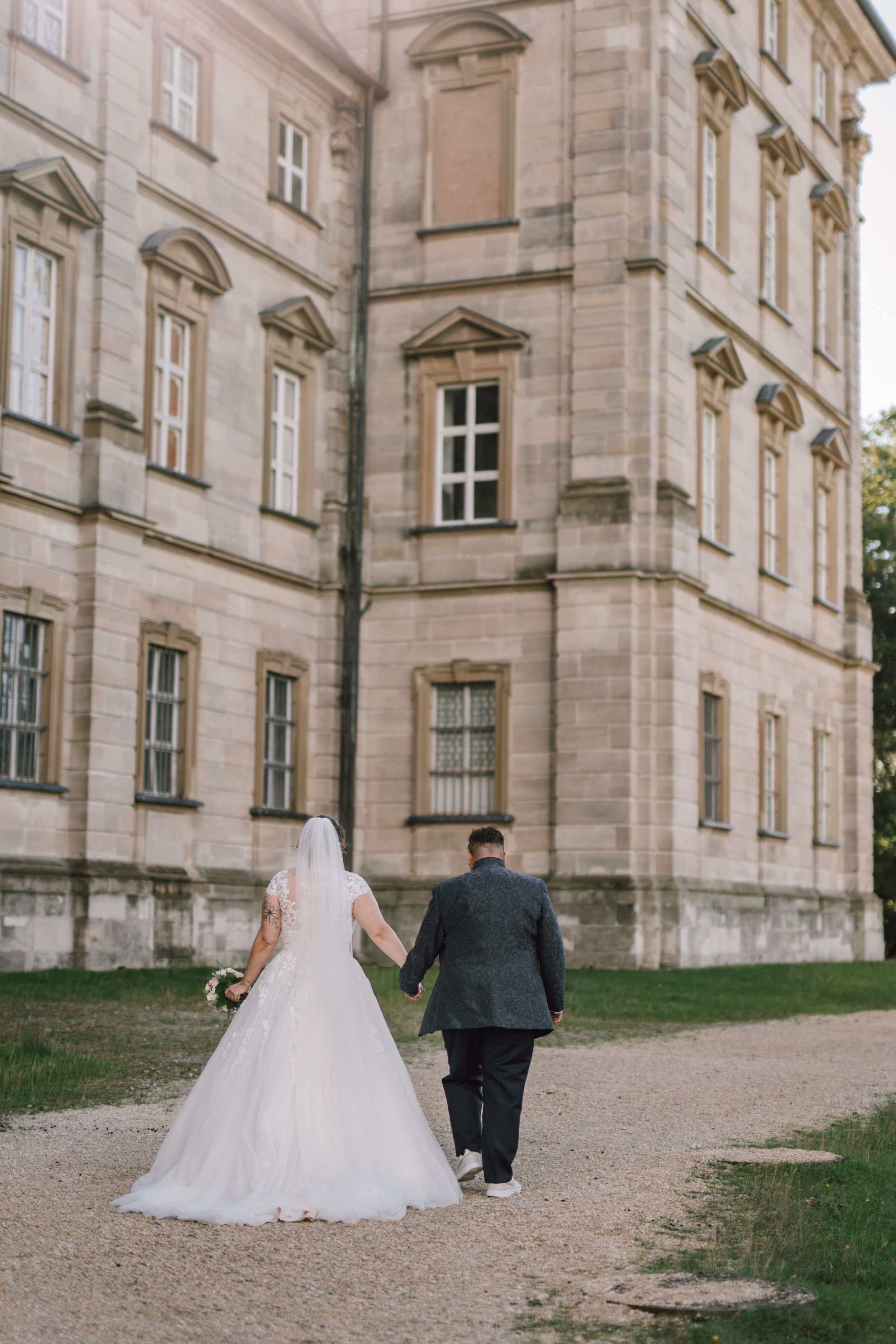 A couple walks along a gravel path beside a grand historic stone building on their wedding day.