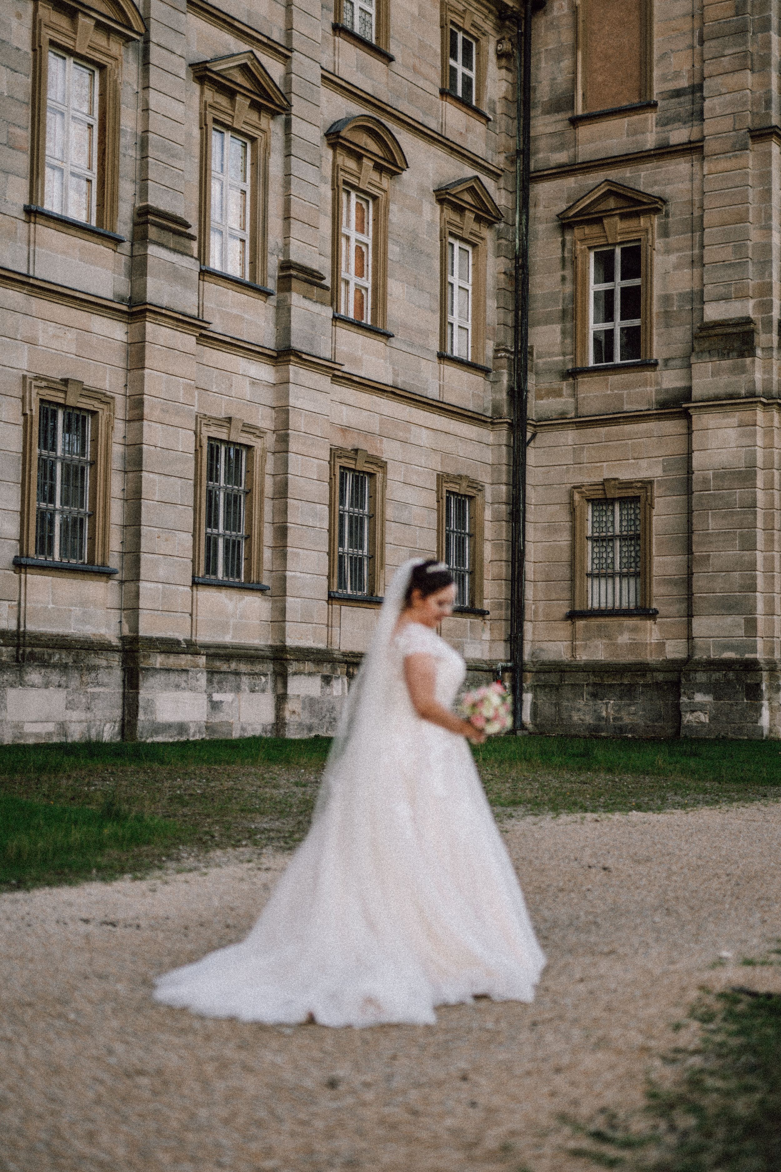 A bride in a flowing white gown walks beside a historic stone building with multiple windows.