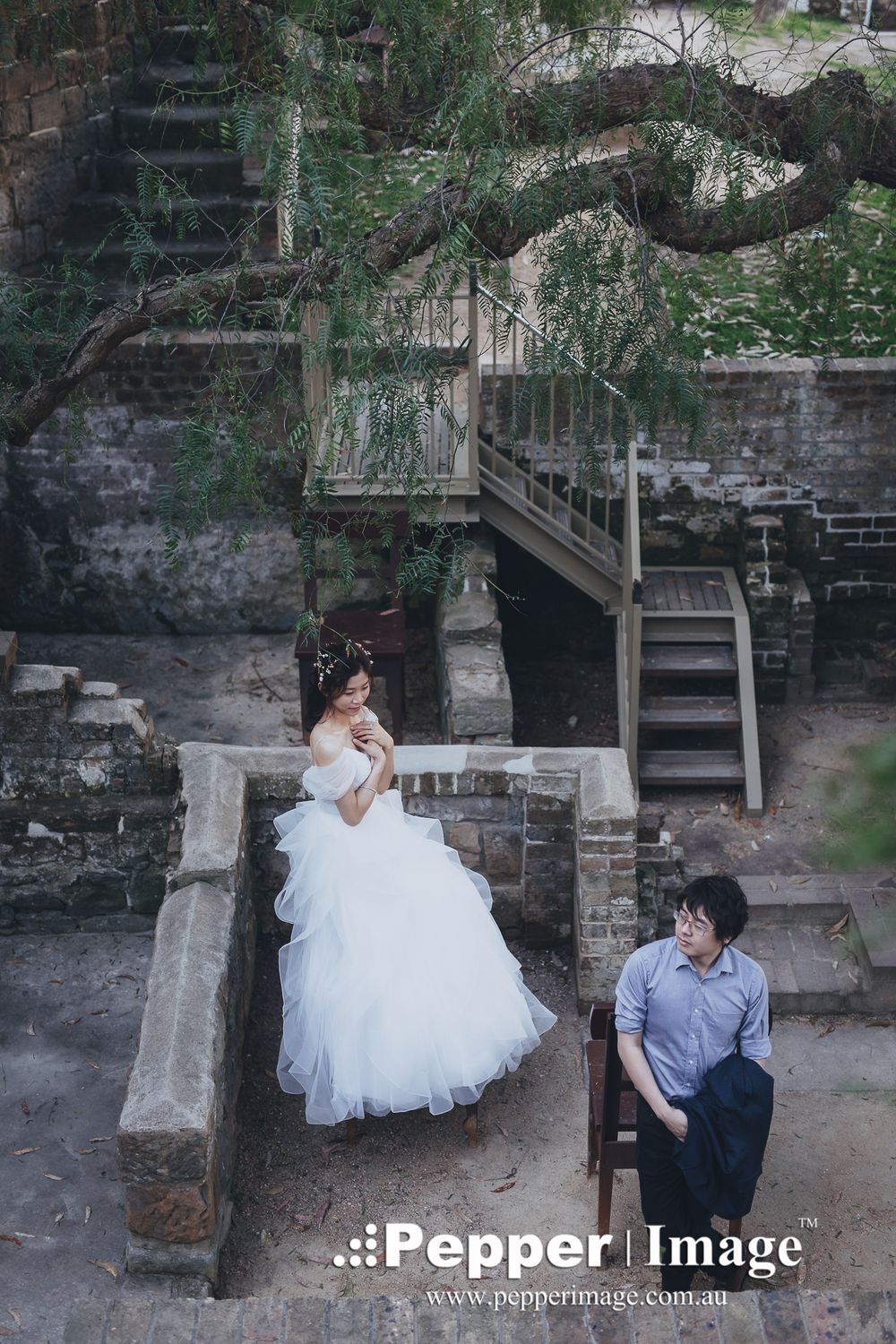 Bride in white ball gown sits on stone steps as groom relaxes nearby in a rustic outdoor setting.