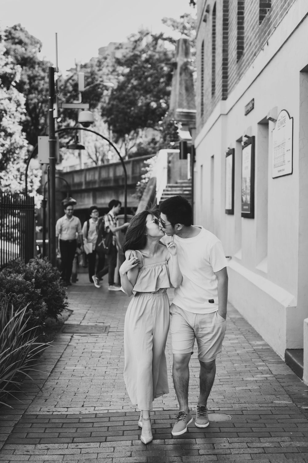 Black and white photo of couple walking and sharing a kiss on city sidewalk.