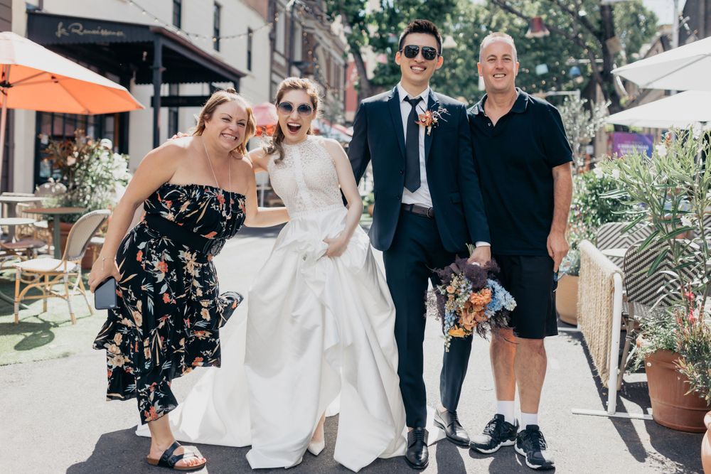Wedding party of four posing on a sunny urban street, bride in white gown with groom in navy suit.