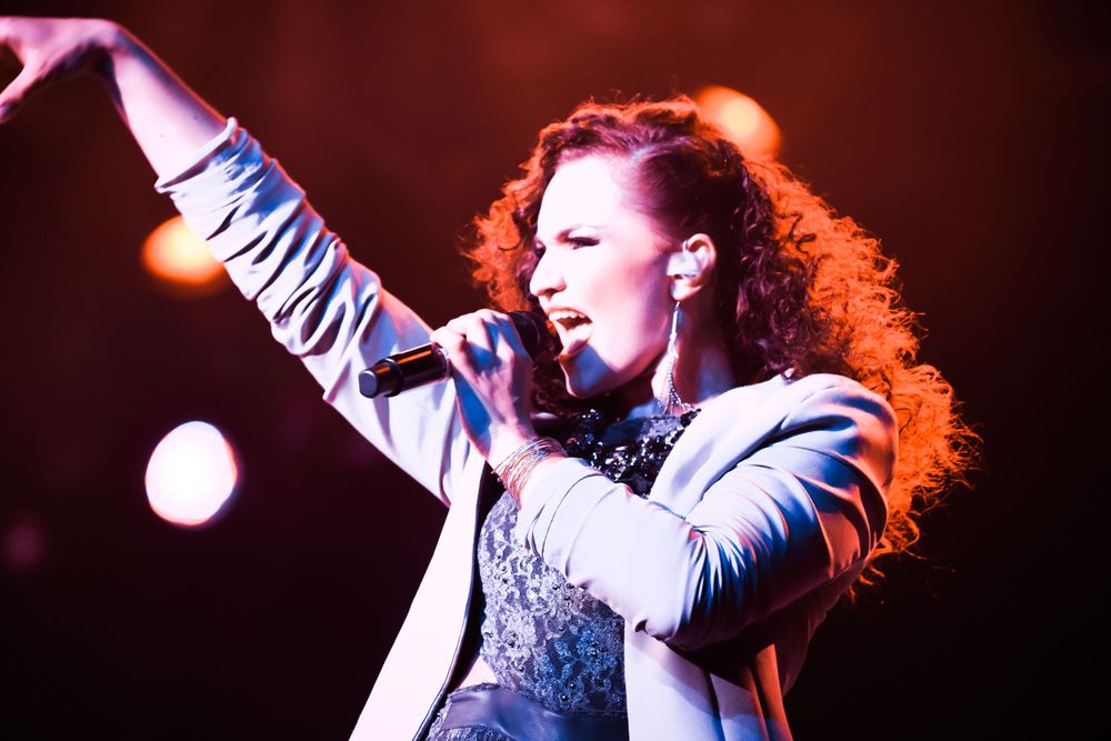 A dynamic performance shot with dramatic lighting showing a singer in a blazer against a dark red background.