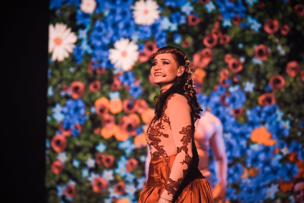 A performer in a vintage-style dress poses against a backdrop of colorful flowers and blue lighting.