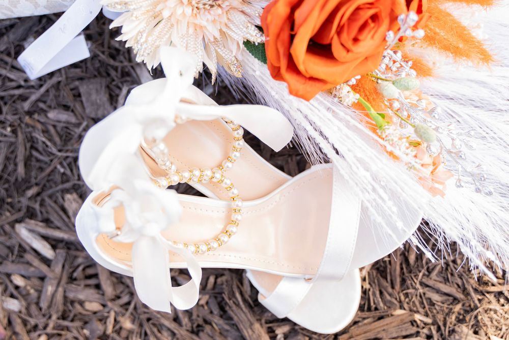 Bridal accessories including pearl-adorned heels and orange rose bouquet arranged on dark mulch.
