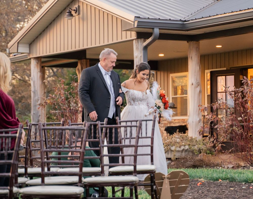 Bride in white gown walks with escort down outdoor wedding aisle surrounded by autumn foliage.