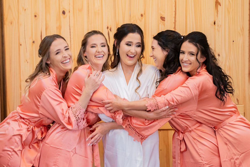 Smiling bride in white robe surrounded by four bridesmaids in coral satin robes before wedding.