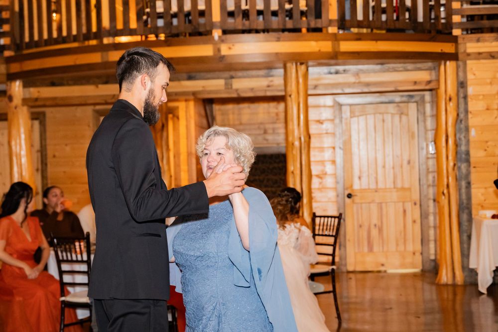 Groom in black tuxedo sharing a tender dance with an older woman at rustic barn wedding reception.