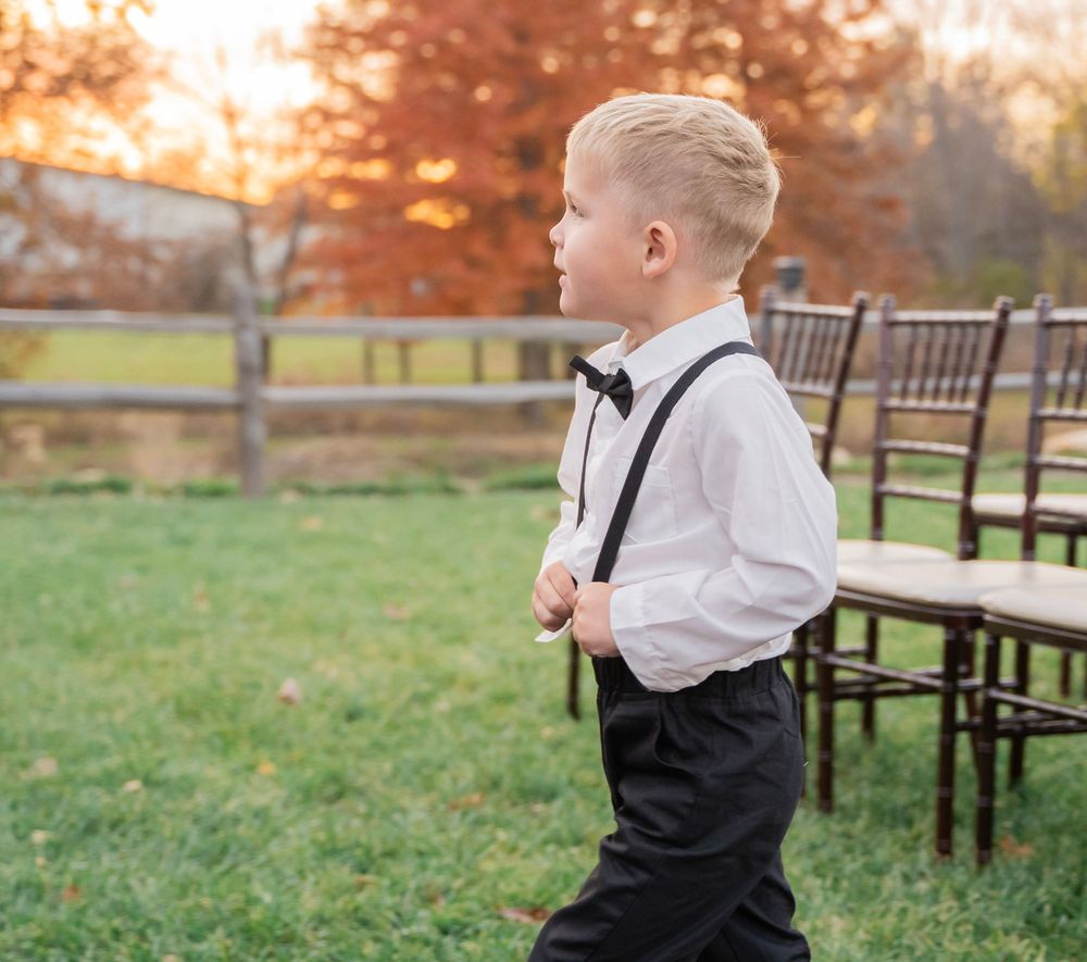Young ring bearer in white shirt, suspenders and bow tie stands on grass at outdoor fall wedding.