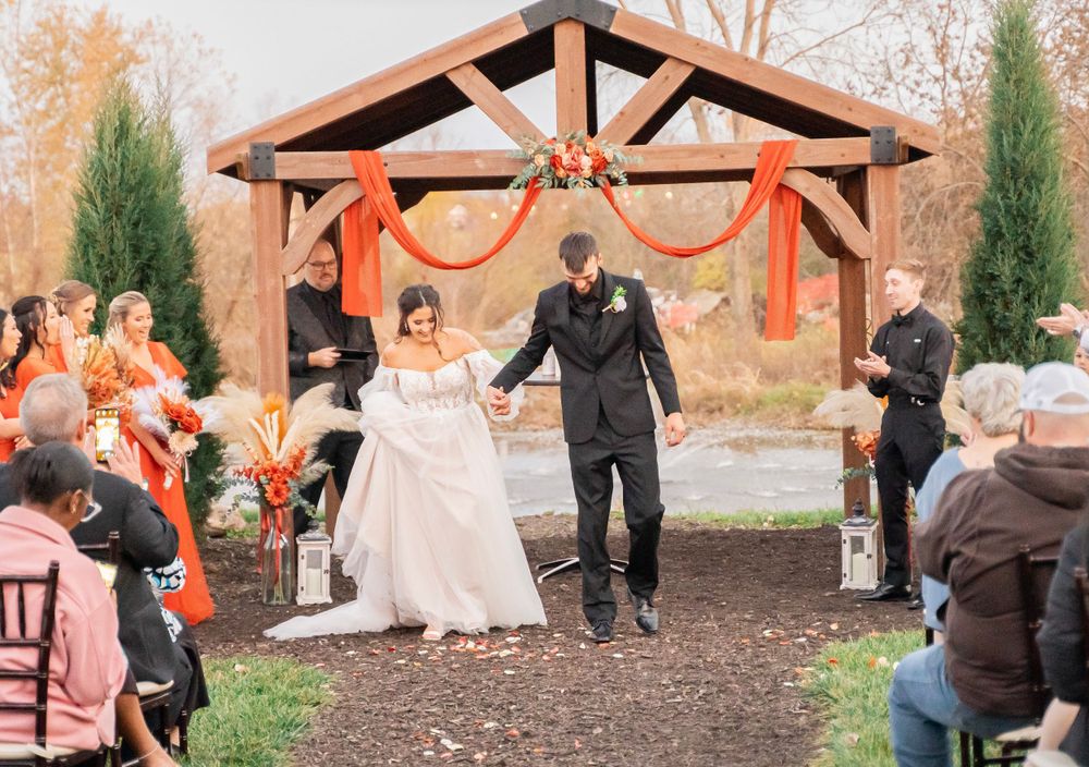 Bride and groom exit outdoor wedding ceremony under a wooden pergola decorated with orange draping.