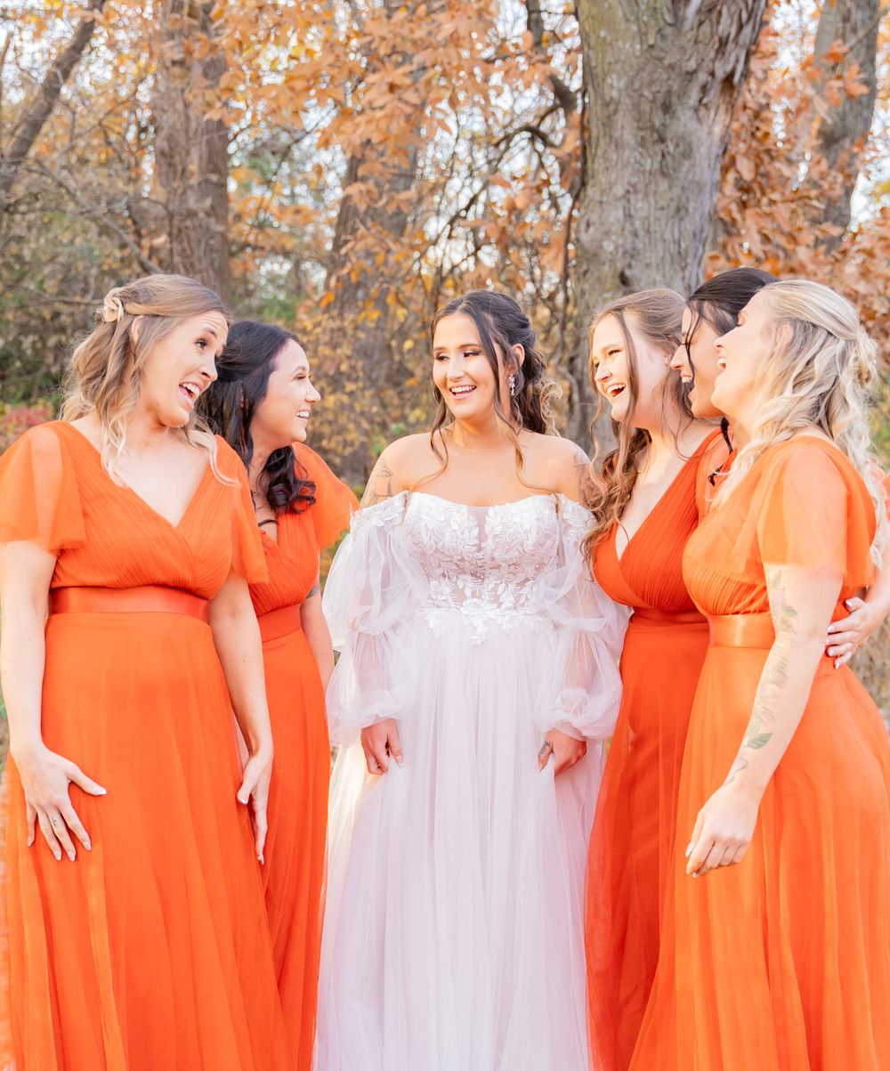 Bride in white off-shoulder gown laughs with four bridesmaids in orange dresses among fall trees.