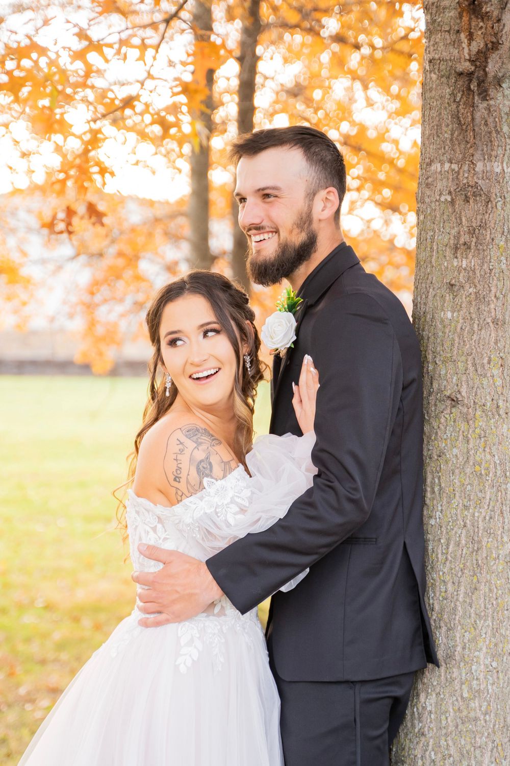 Laughing bride with tattoo leans against groom in black suit by tree with fall foliage.