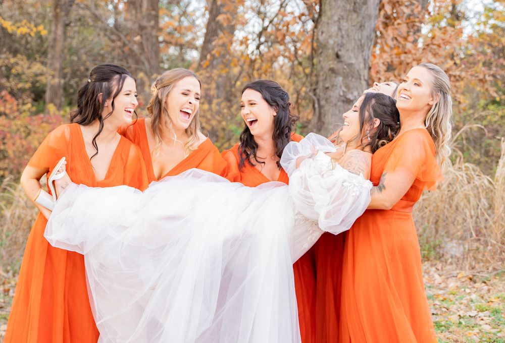 Bridesmaids in orange gowns joyfully lift bride in white dress during autumn outdoor wedding.