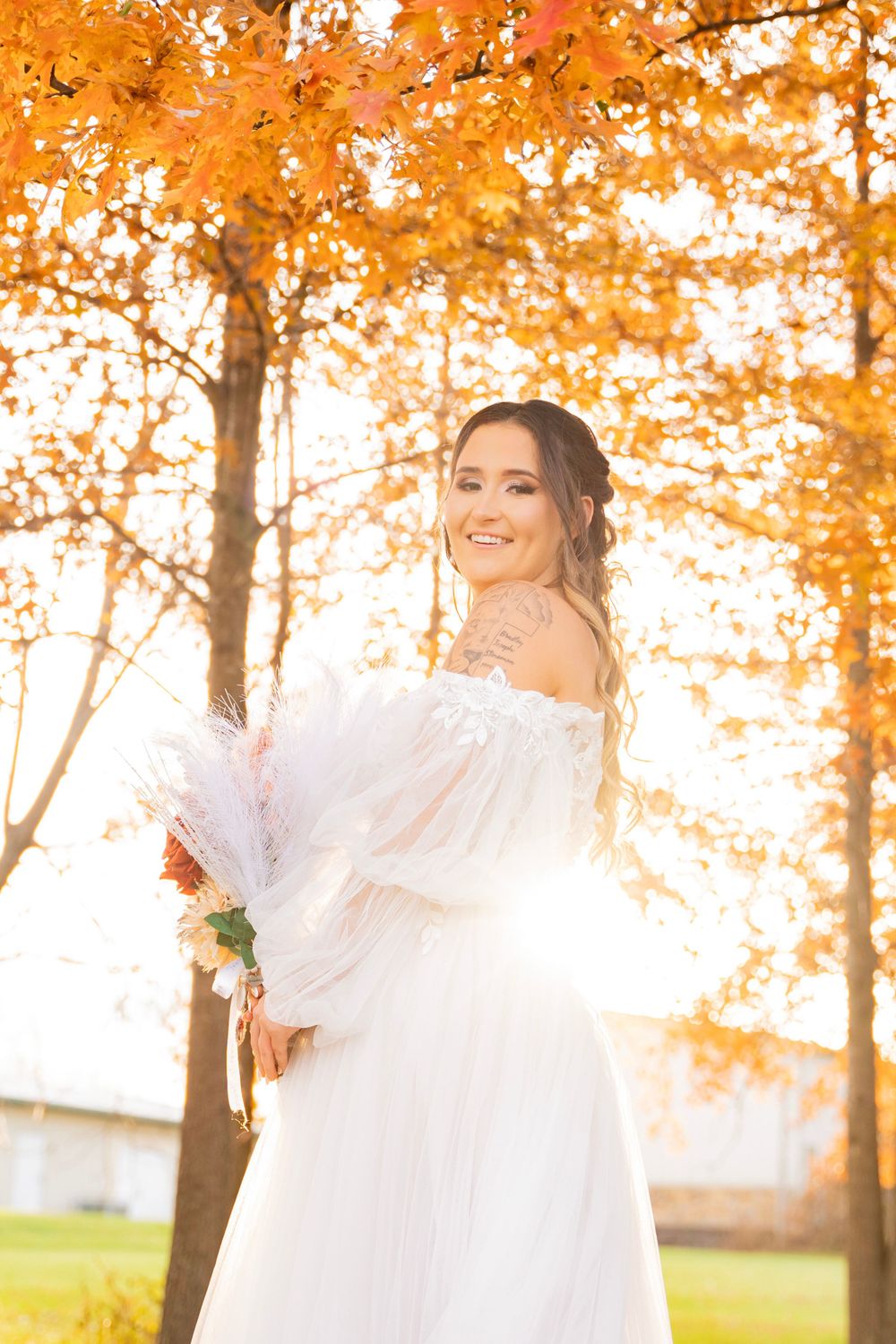 Smiling bride in white off-shoulder gown holds bouquet under golden autumn trees in sunlight.