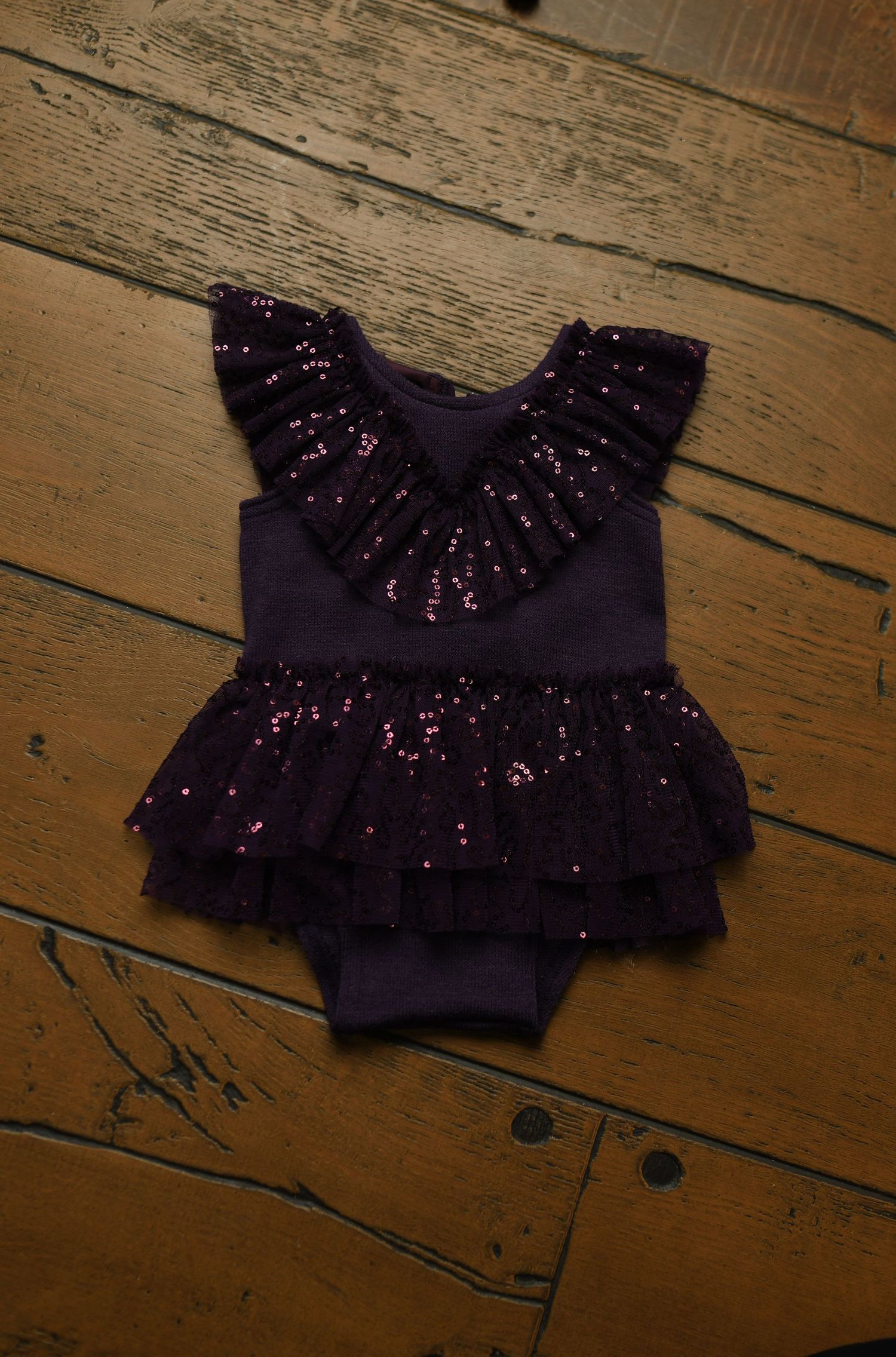 Purple sparkly baby romper with ruffle detail photographed on wooden floor.