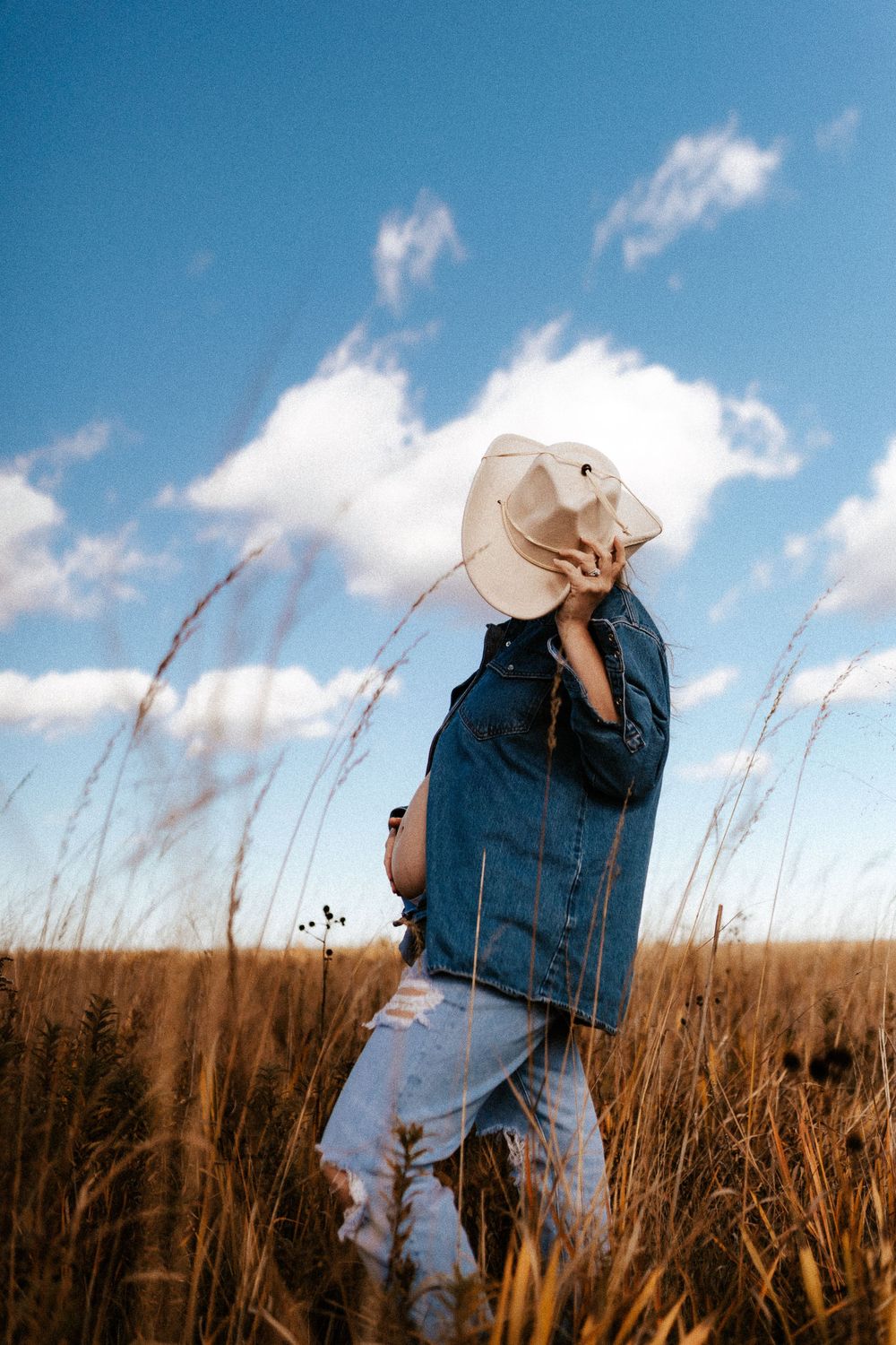 Figure in denim jacket and cowboy hat standing in golden field against blue sky with wispy clouds.