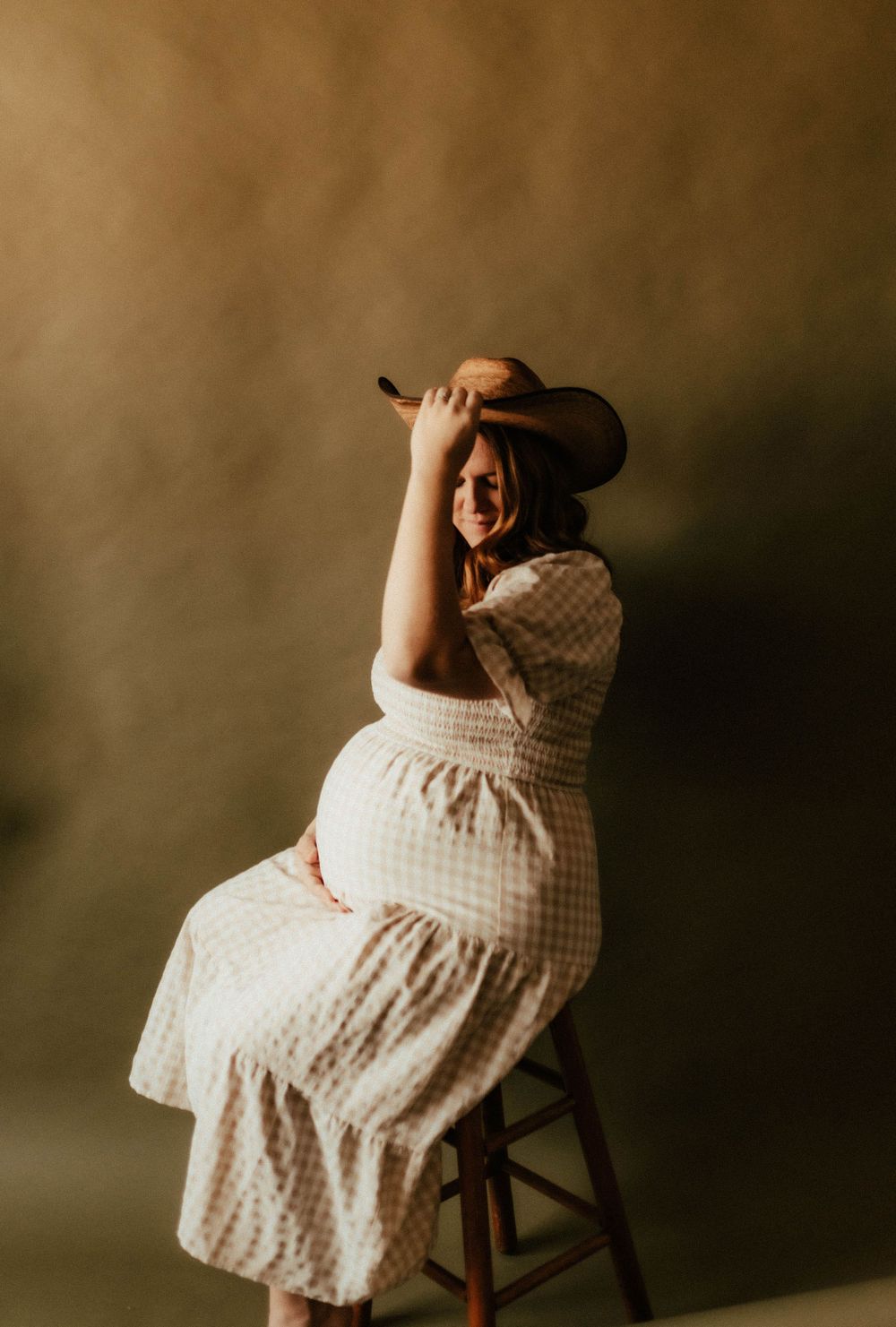 Pregnant woman in gingham dress poses on stool wearing cowboy hat against moody backdrop.