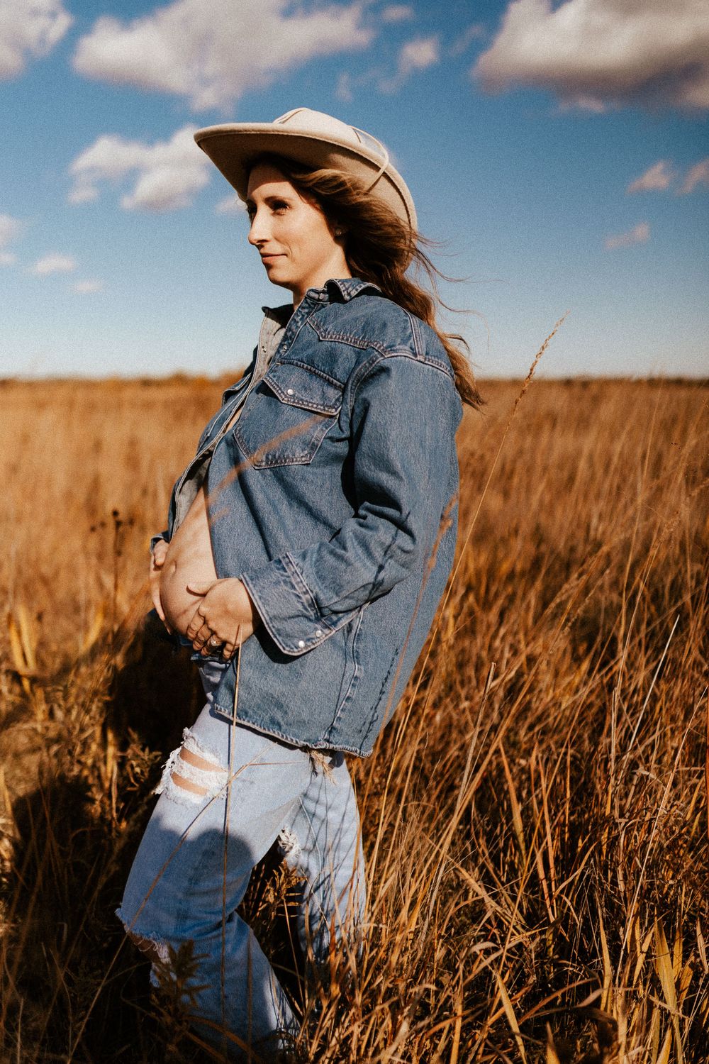 Maternity portrait in denim jacket and cowboy hat standing in golden field during sunset.