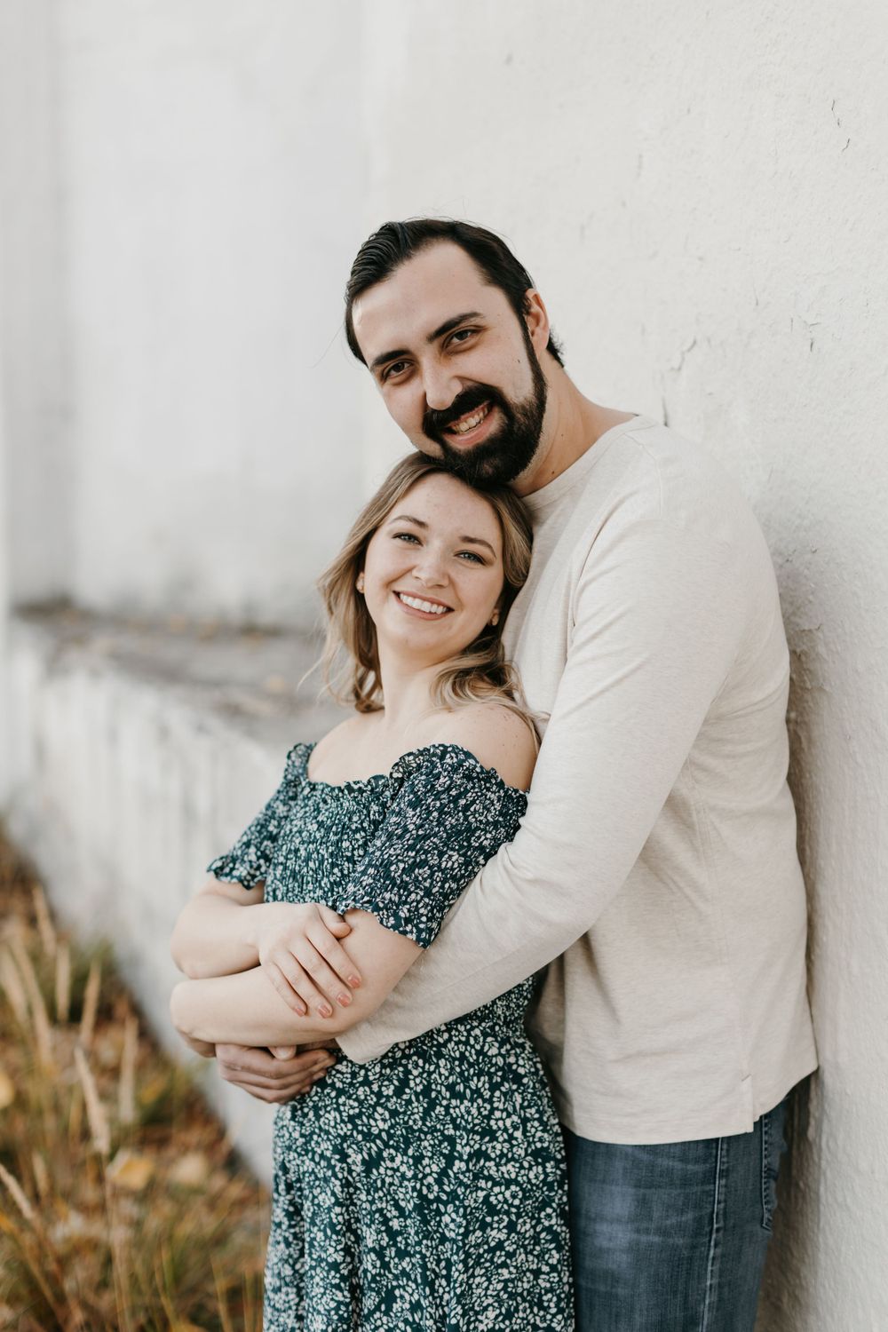 Joyful couple hugging against a white wall, man in cream sweater embracing woman in off-shoulder floral dress.