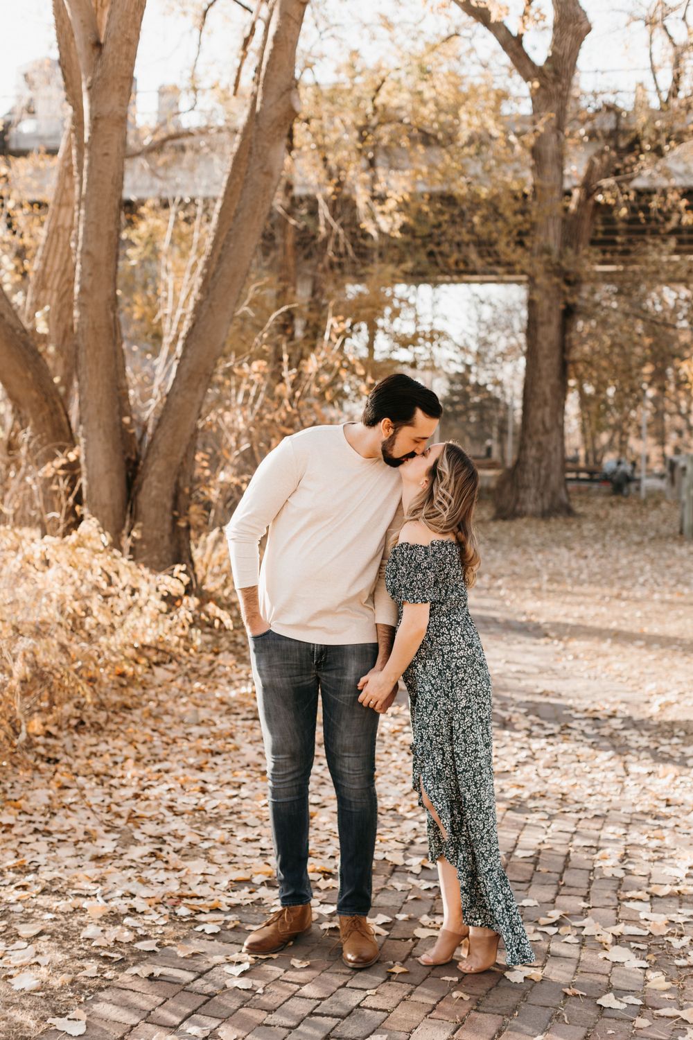 Couple sharing a romantic kiss while holding hands on a brick path with golden autumn trees in the background.