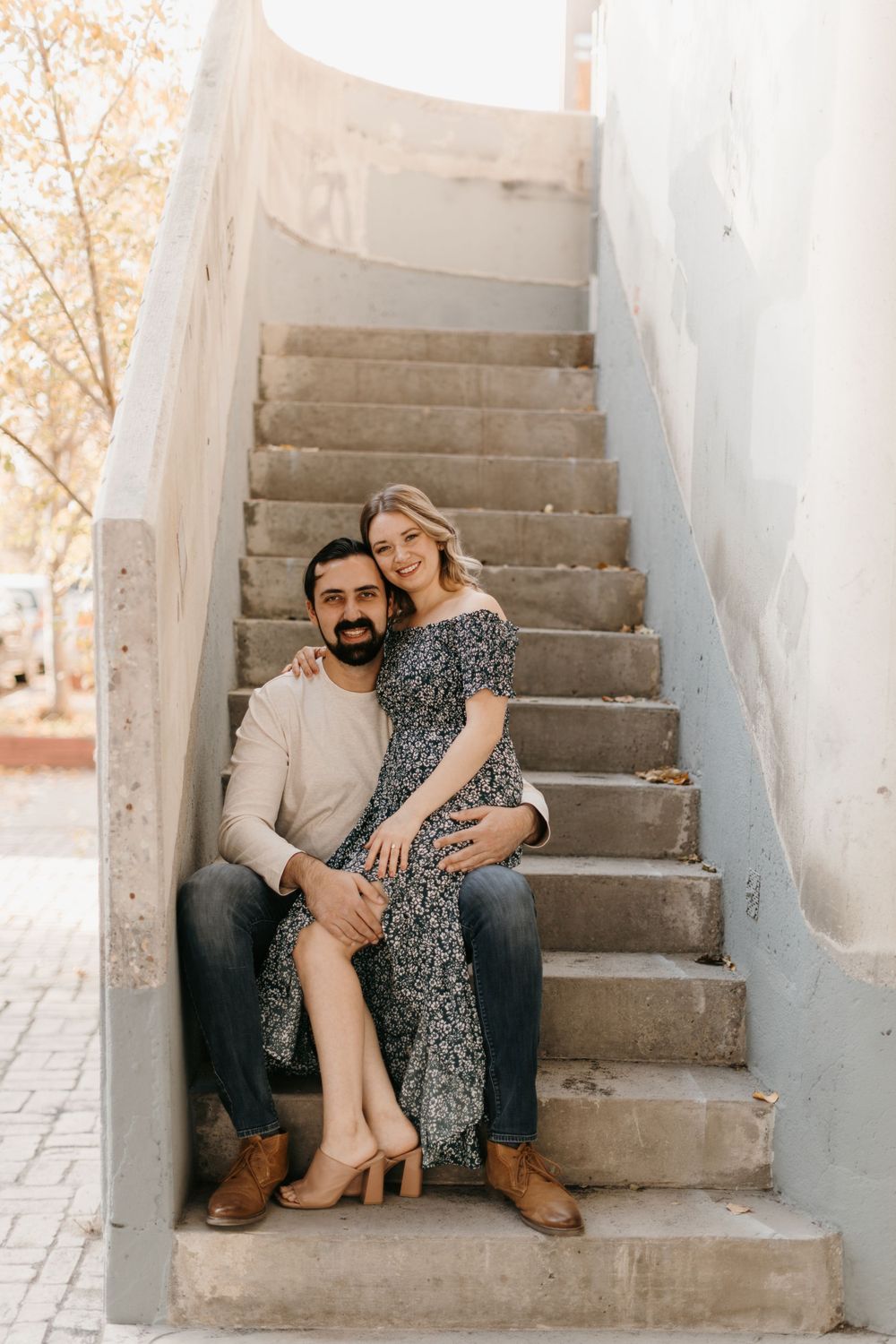 A smiling couple poses together on concrete stairs, woman in floral dress leaning against man.