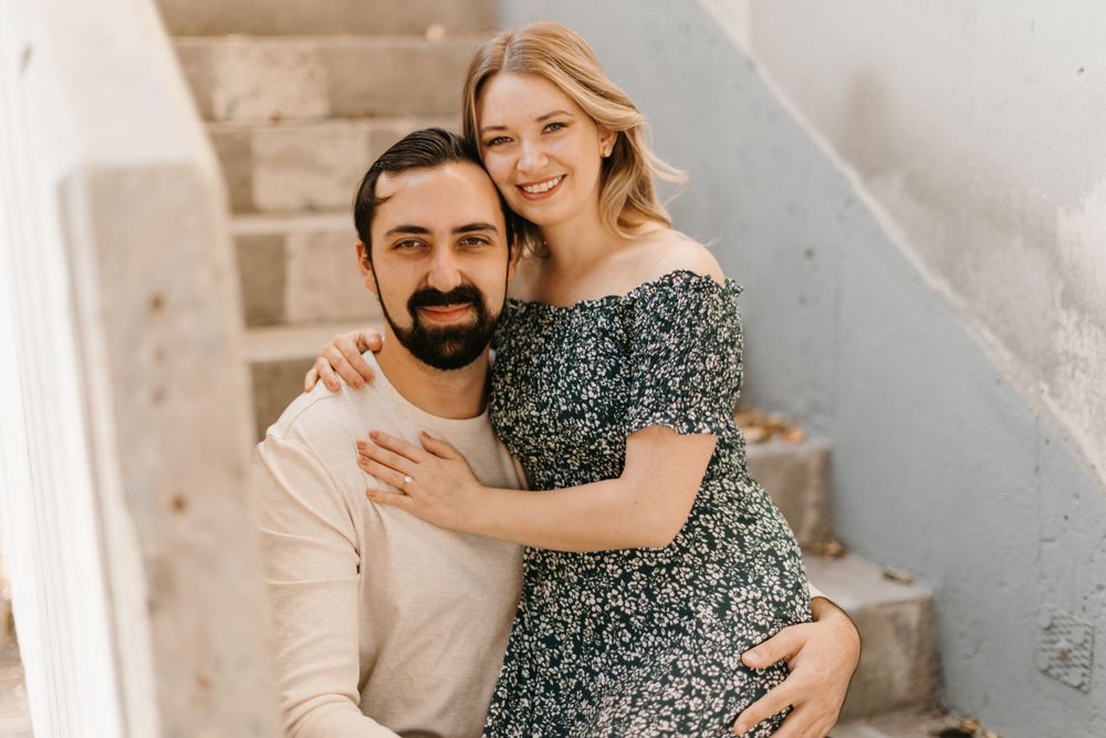 Smiling couple embracing on white stone steps, woman in floral dress hugging bearded man in cream sweater.