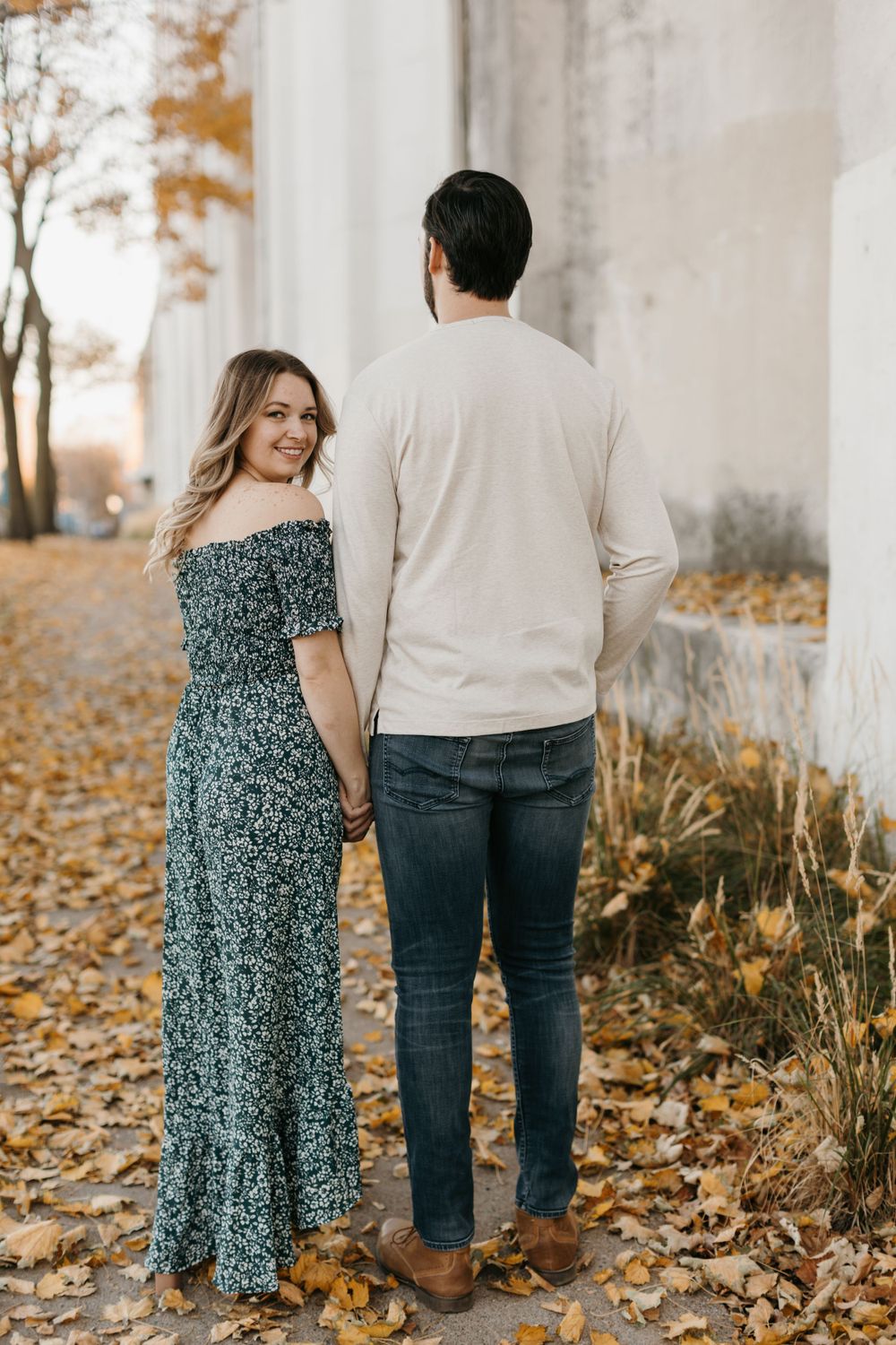 Couple holding hands walking past white pillars on a leaf-covered autumn path.