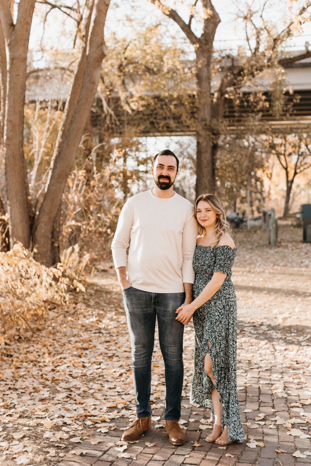 Couple standing together on brick path surrounded by fall foliage, man in cream sweater and woman in floral dress.