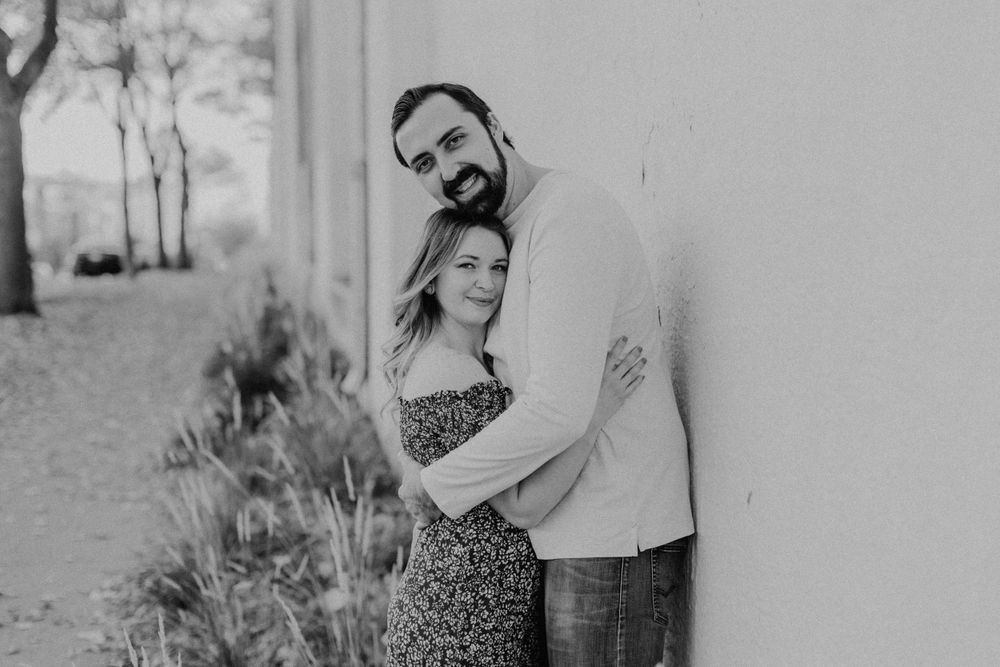 Black and white photo of couple embracing tenderly against a white wall with soft natural lighting.