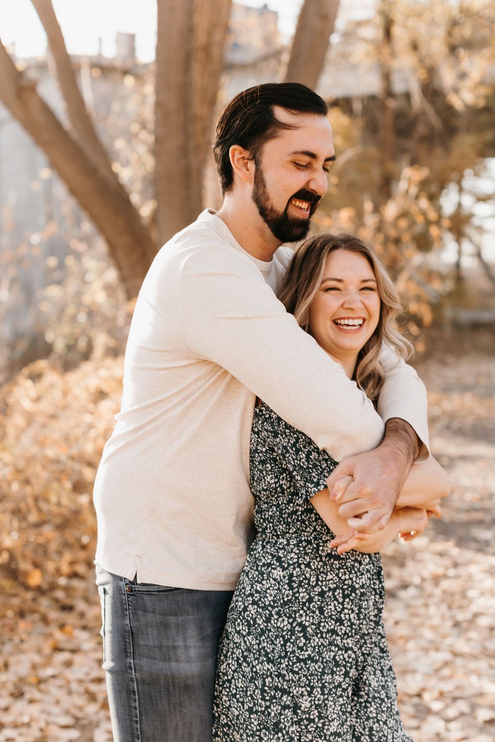 Couple sharing a joyful embrace outdoors in golden autumn light, woman laughing as man hugs her from behind.