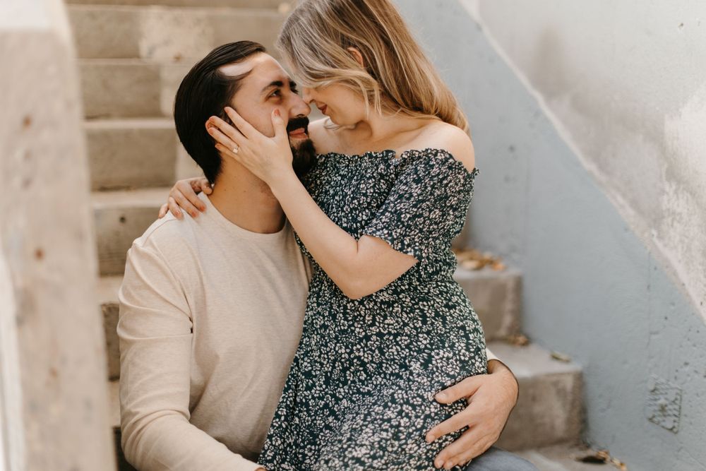 A couple leans in for a kiss on concrete stairs, woman cradling mans face with both hands.