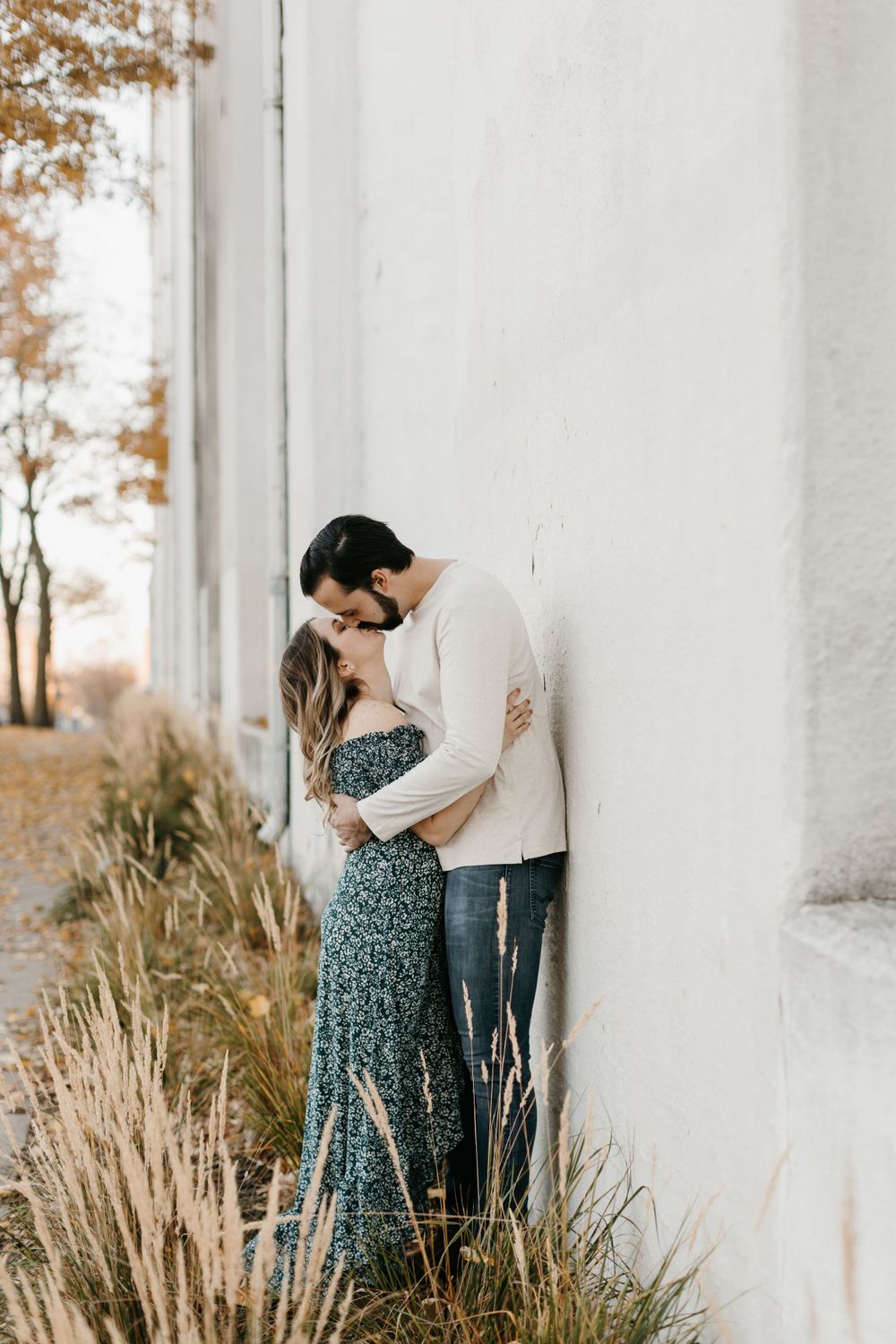 Romantic couple sharing an intimate moment leaning against a white wall with autumn foliage in background.