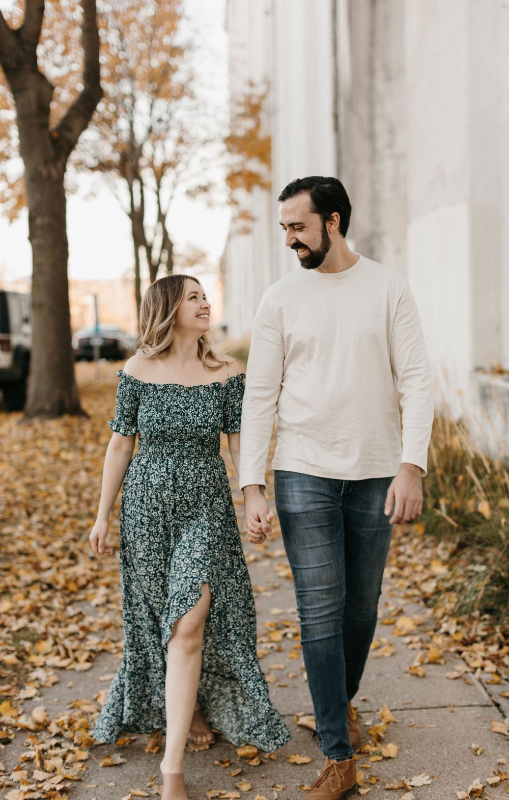 Smiling couple strolling hand in hand among fallen autumn leaves, woman in green floral maxi dress, man in cream sweater.