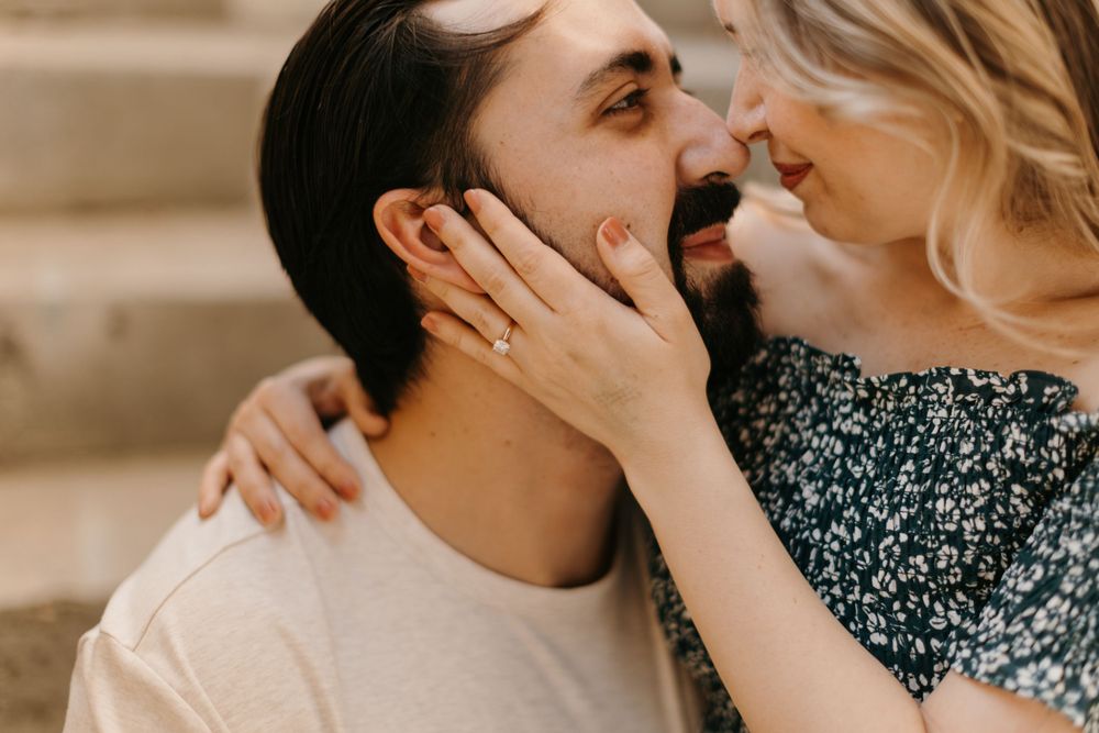 A close-up of a couple almost kissing, woman touching mans bearded face showing an engagement ring.