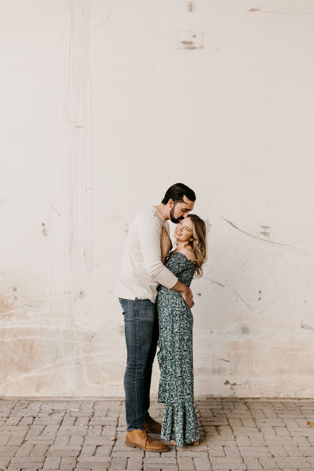 Romantic couple nearly kissing while embracing against a rustic white wall on a brick pathway.