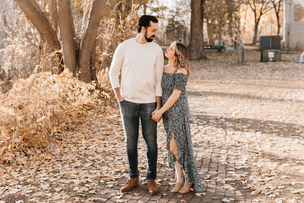 Couple smiling at each other while holding hands on a brick path surrounded by golden autumn leaves and trees.